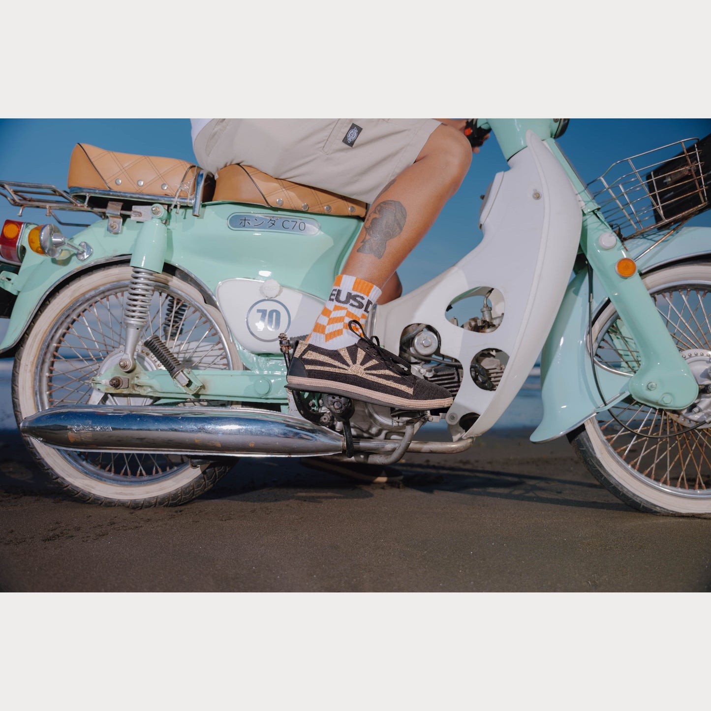 A person sitting on a light blue motorbike wearing hemp sneakers with a sunset design on the upper part.